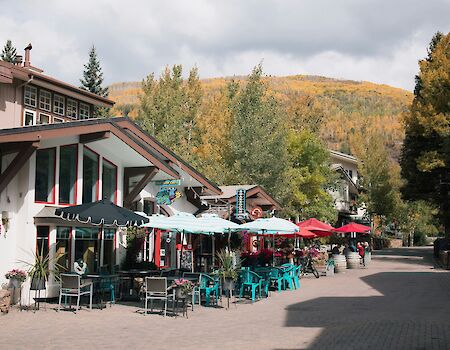 A quaint mountain village street with cafes and outdoor seating under colorful umbrellas, nestled among trees with yellowing hills in the background.