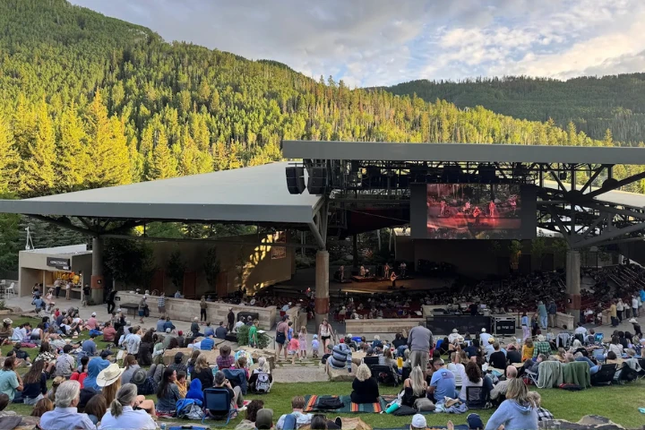 An outdoor amphitheater sits in a grassy field with a crowd watching a performance on stage, surrounded by forested hills under a blue sky.