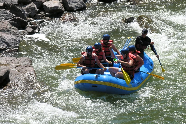 A group of six people wearing life jackets and helmets navigate a blue inflatable raft with paddles down a rocky, white-water river.