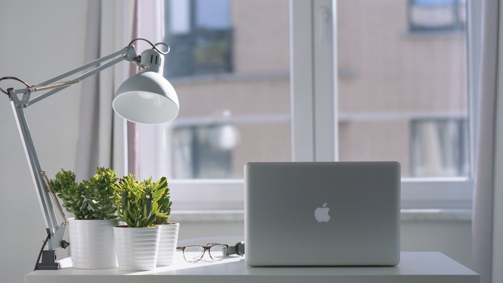 A minimalistic desk setup featuring a laptop, potted plants, a desk lamp, and a pair of glasses, situated in front of a window with curtains.