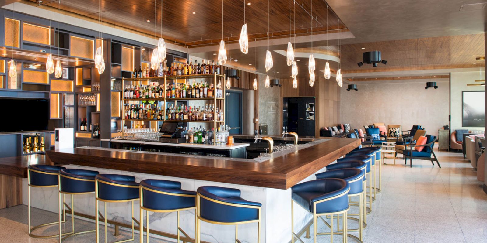 This image shows a modern bar with seating, shelves of liquor bottles, hanging lights, and a lounge area with comfortable seating in the background.