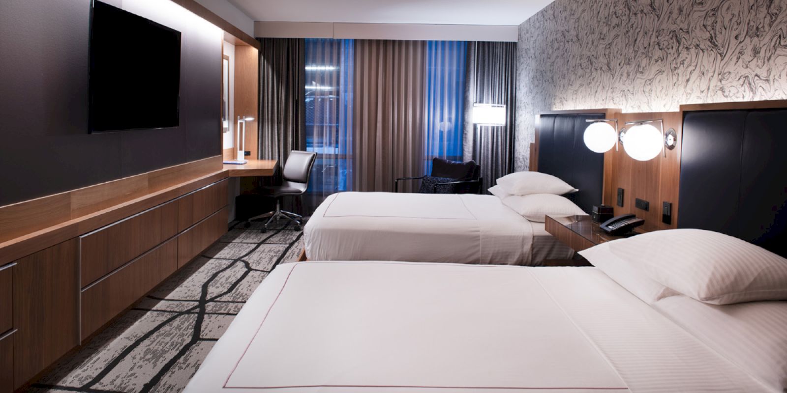 A modern hotel room with two double beds, a wall-mounted TV, a desk with a chair, and a large window with curtains.