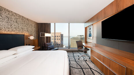The image shows a modern hotel room with a large bed, a desk, a chair, a wall-mounted TV, and a window with a city view in the background.