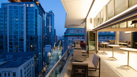 A modern balcony with tables and chairs overlooks a cityscape at dusk, featuring tall buildings and a bustling street below.