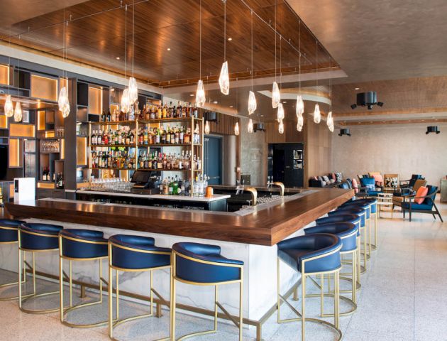A modern bar with a wooden countertop, blue cushioned stools, and hanging lights, featuring a wide array of bottles behind the counter.