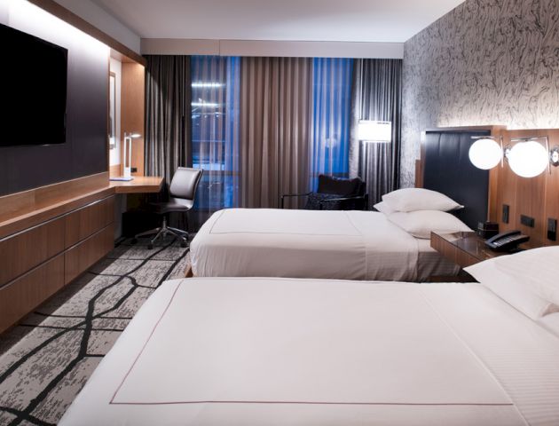 A modern hotel room with two double beds, a flat-screen TV, a work desk with a chair, and contemporary decor on the walls and floor.