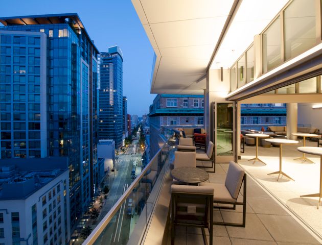 An outdoor terrace with tables and chairs overlooks a lit urban street lined with modern high-rise buildings at dusk.