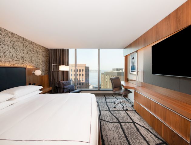 The image shows a modern hotel room with a large bed, a TV, a desk and chair, a cityscape view through floor-to-ceiling windows, and contemporary decor.