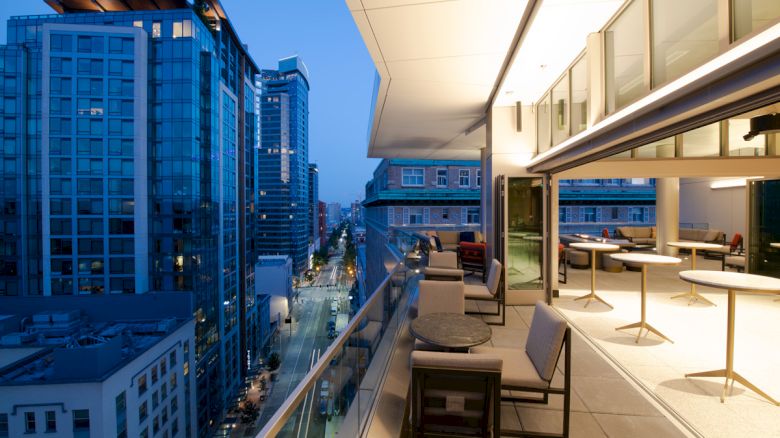 A modern cityscape with tall buildings at dusk, viewed from a balcony with tables, chairs, and a seating area along the railing.