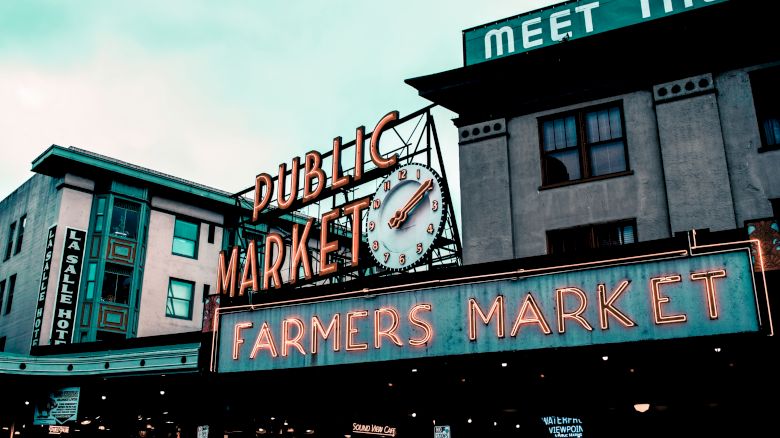 The image shows a neon sign reading "Public Market" and "Farmers Market" with a clock above it, set against urban buildings on a cloudy day.