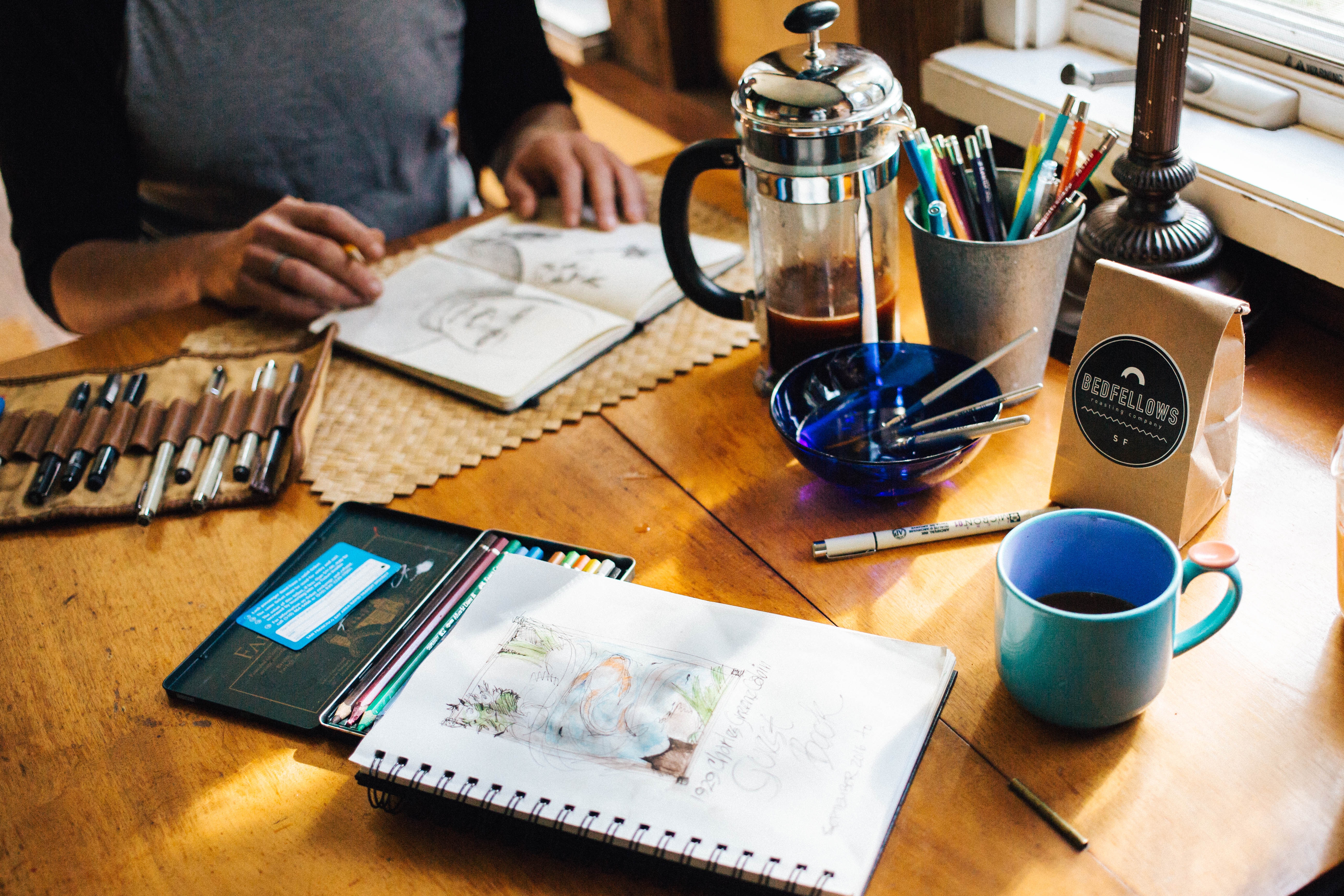 A person is drawing in a notebook on a wooden table with various art supplies, including pencils and pens, and a cup of coffee.